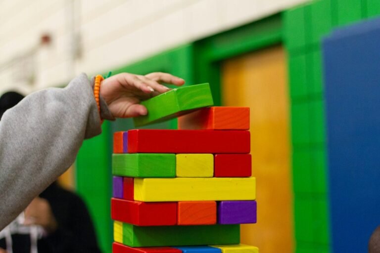 A child's hand adding a green wooden block to a colorful tower.